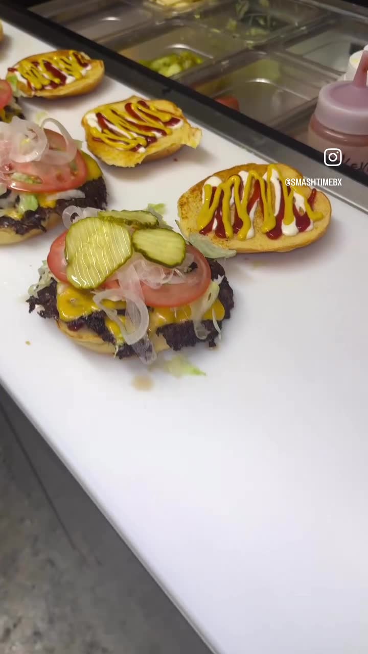 Smash patties on the board being dressed with pickles, tomato, onion and mustard