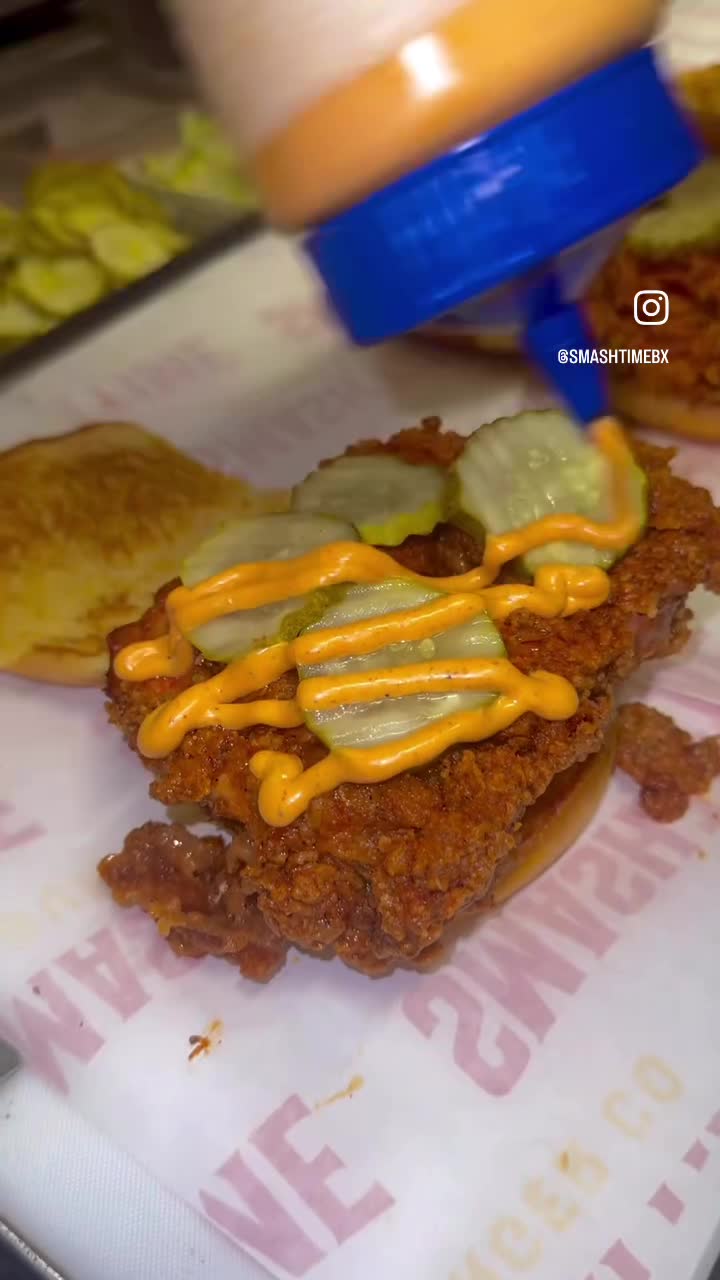 A chicken sandwich with pickles being finished with a drizzle of sauce